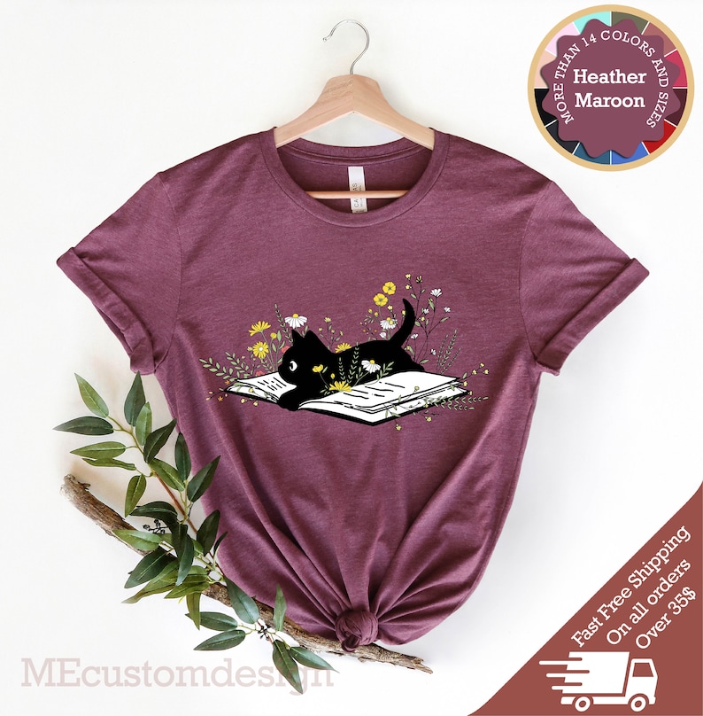 May include: A heather maroon t-shirt with a black cat reading a book surrounded by flowers. The text "More Than 14 Colors and Sizes" is on a circle with a variety of colored circles around it. The text "Heather Maroon" is in the center of the circle.