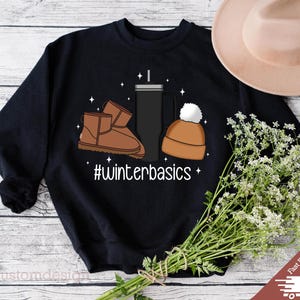 May include: Black sweatshirt with a graphic design of a brown winter boot, a brown beanie, and a black travel mug. The text "#winterbasics" is printed below the graphic.