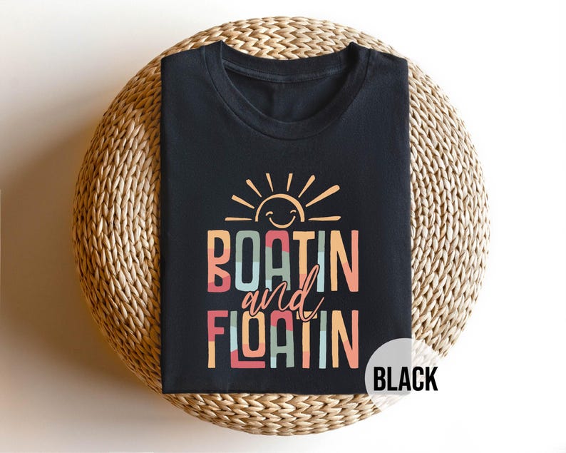 May include: Black t-shirt with the words "BOATIN and FLOATIN" in colorful block letters, topped with a smiling sun graphic. The shirt is displayed on a woven, circular surface. The word "BLACK" is in a white circle.