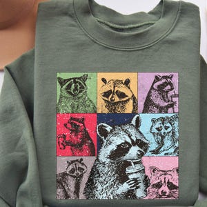 May include: A green sweatshirt with a colorful graphic of raccoons drinking from cups. The raccoons are in a grid pattern with each raccoon in a different colored square.