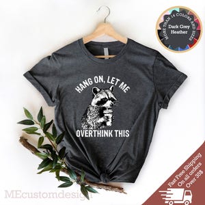 May include: Dark heather grey t-shirt with a raccoon graphic and the text "HANG ON, LET ME OVERTHINK THIS".