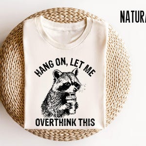 May include: A white t-shirt with a black and white illustration of a raccoon holding a cup of coffee. The text "Hang on, let me overthink this" is printed on the shirt.