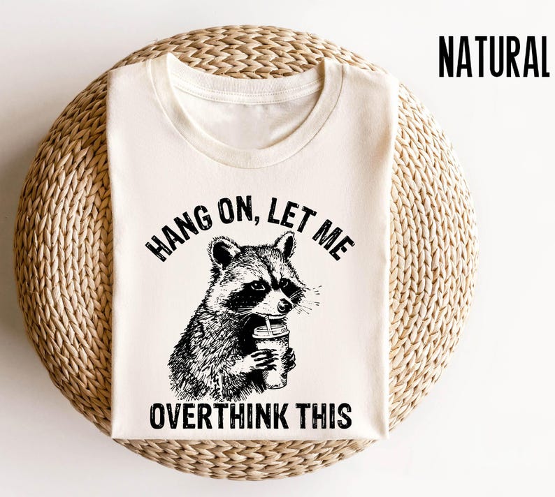 May include: A white t-shirt with a black and white illustration of a raccoon holding a cup of coffee. The text "Hang on, let me overthink this" is printed on the shirt.