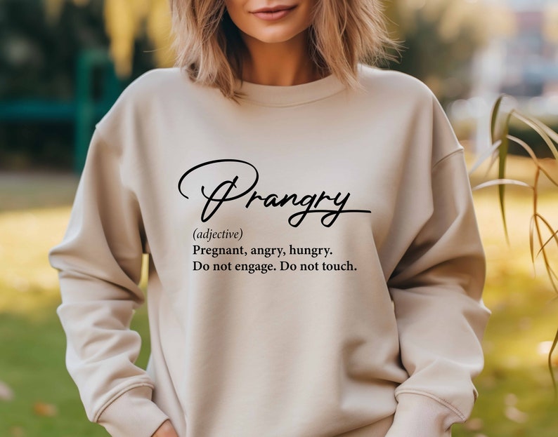 May include: A beige sweatshirt with the word "Prangry" written in black cursive script. The word is defined as "Pregnant, angry, hungry. Do not engage. Do not touch."