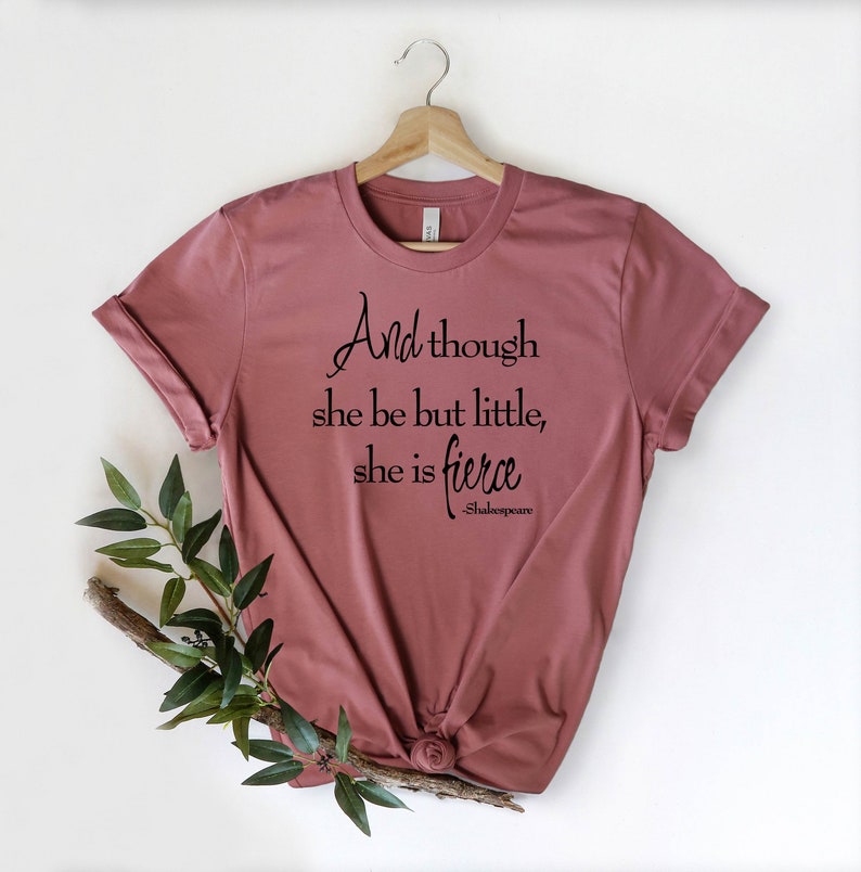 May include: Dusty rose t-shirt with the quote "And though she be but little, she is fierce" in black script. The Shakespeare quote is displayed on a folded t-shirt, with a wooden hanger and greenery.