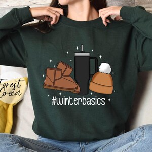 May include: Forest green sweatshirt with a graphic design of a pair of brown Ugg boots, a black travel mug, and a brown beanie with a white pom pom. The text "#winterbasics" is printed below the graphic.