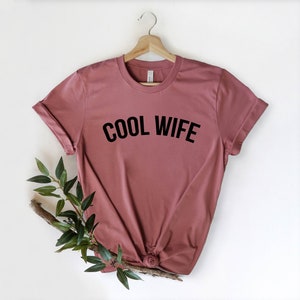 Puede incluir: Una camiseta color malva con el texto "COOL WIFE" impreso en negro.