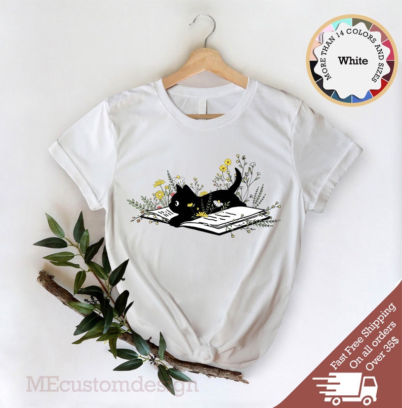 May include: White t-shirt with a black cat reading a book surrounded by yellow flowers. The cat is looking at the book. The text "MORE THAN 14 COLORS AND SIZES" is in a circle above the cat.