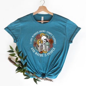 Pu&ograve; includere: Una maglietta blu turchese con una grafica circolare bianca che presenta uno scheletro che tiene una tazza di caff&egrave; circondato da fiori colorati. Il testo recita "Mi piacciono gli show di omicidi, i vestiti comodi e forse 3 persone."