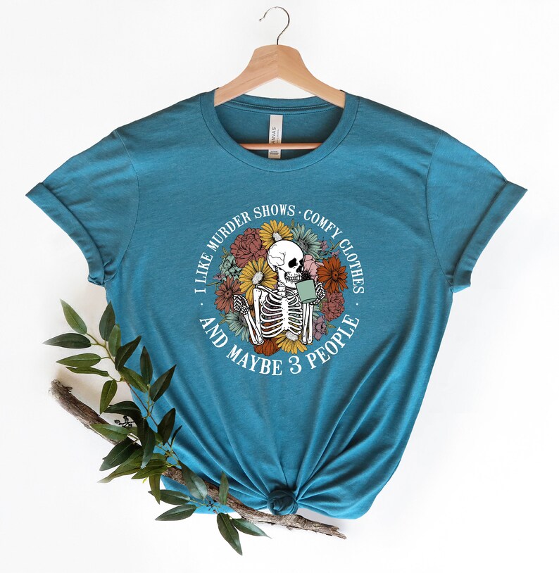 Pu&ograve; includere: Una maglietta blu turchese con una grafica circolare bianca che presenta uno scheletro che tiene una tazza di caff&egrave; circondato da fiori colorati. Il testo recita "Mi piacciono gli show di omicidi, i vestiti comodi e forse 3 persone."