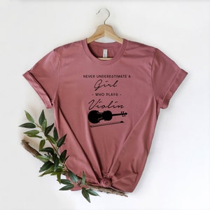 May include: A mauve t-shirt with the text "Never underestimate a girl who plays violin" and a black silhouette of a violin with a bow.