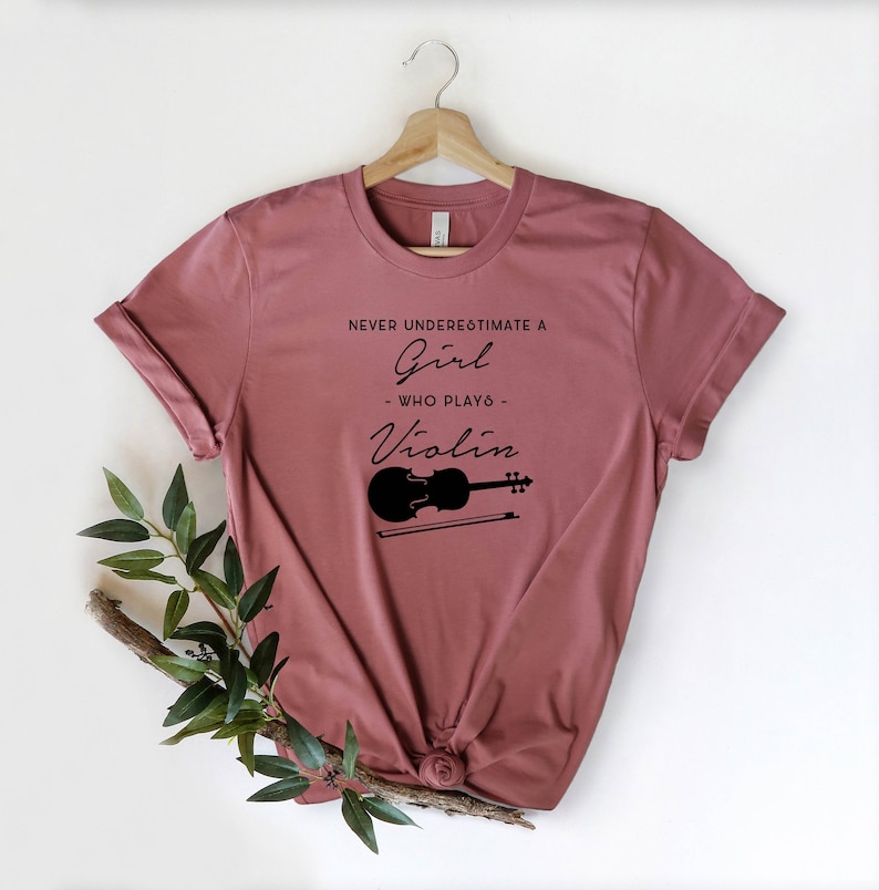 May include: A mauve t-shirt with the text "Never underestimate a girl who plays violin" and a black silhouette of a violin with a bow.