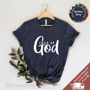 May include: A navy blue t-shirt with the text "CHILD OF GOD" in white lettering. The shirt is hanging on a wooden hanger against a white background.