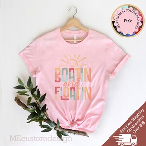 May include: A light pink t-shirt with the words "BOATING and FLOATIN" in colorful letters, with a smiling sun graphic above. The shirt is displayed on a wooden hanger. A circular graphic indicates the availability of multiple colors and sizes.