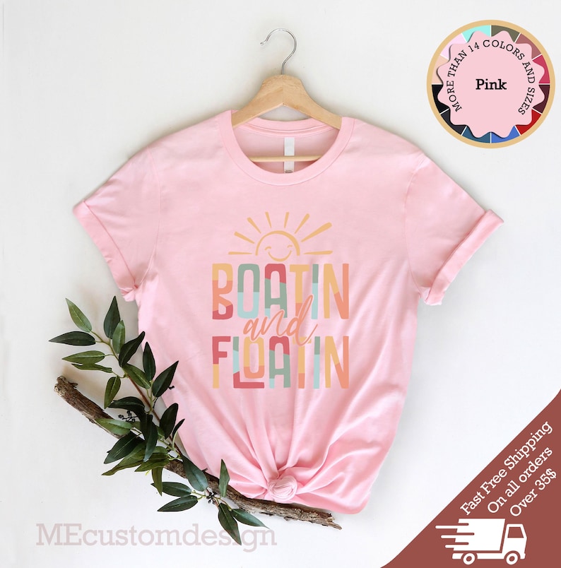 May include: A light pink t-shirt with the words "BOATING and FLOATIN" in colorful letters, with a smiling sun graphic above. The shirt is displayed on a wooden hanger. A circular graphic indicates the availability of multiple colors and sizes.