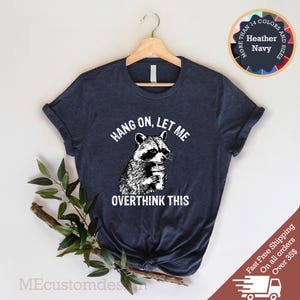 May include: A navy blue t-shirt with a white graphic of a raccoon holding a cup of coffee. The text "Hang on, let me overthink this" is printed below the raccoon.