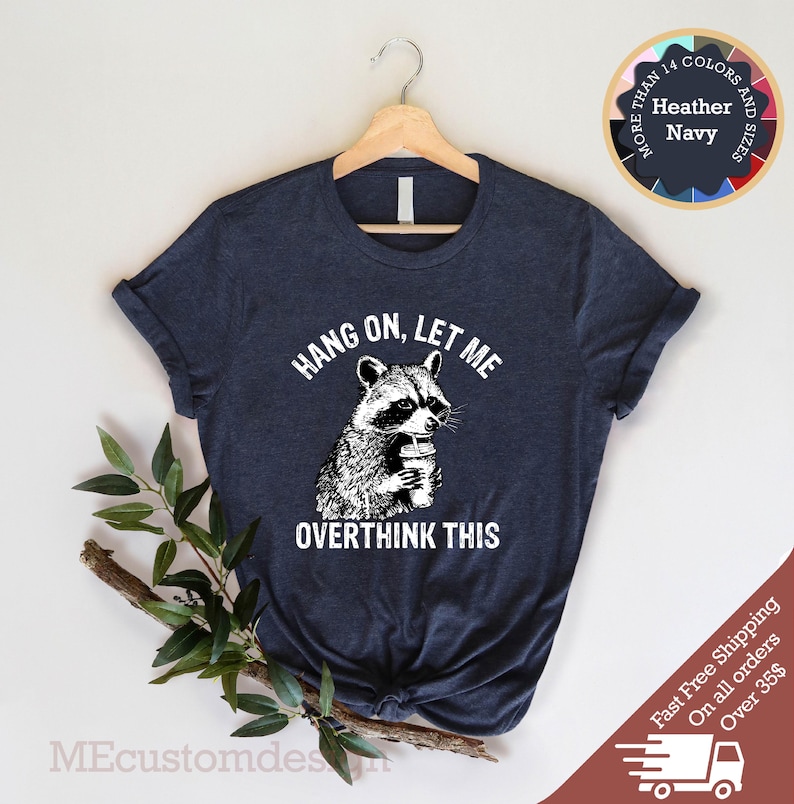 May include: A navy blue t-shirt with a white graphic of a raccoon holding a cup of coffee. The text "Hang on, let me overthink this" is printed below the raccoon.
