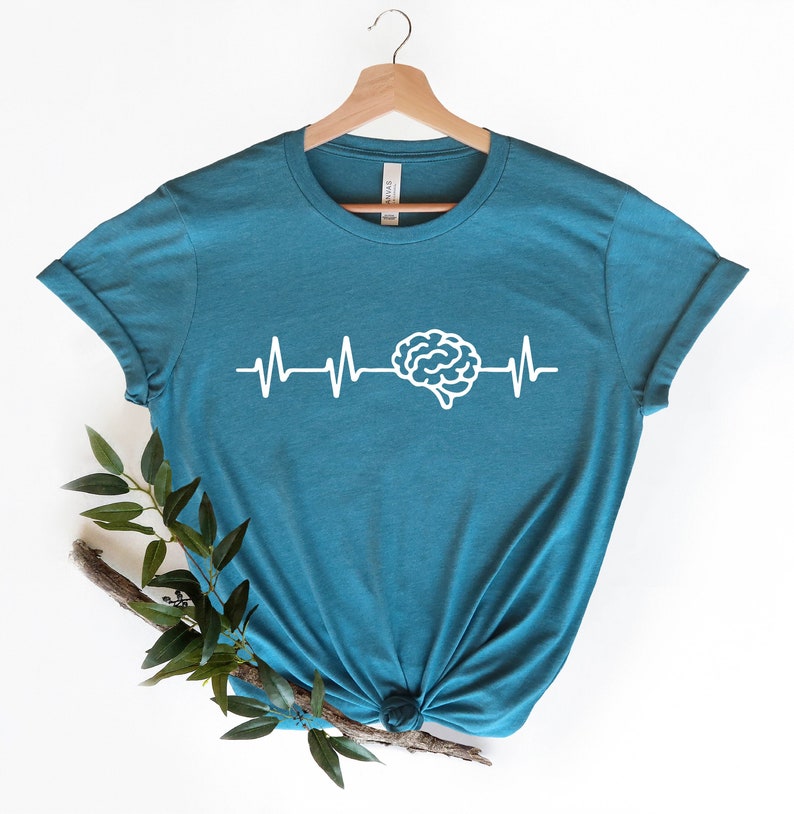 Op de afbeelding: Tealblauw t-shirt met een witte hersengrafiek en een witte hartslaglijn. Het shirt is gevouwen en wordt getoond op een hanger.
