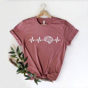 Op de afbeelding: Een maronkleurig t-shirt met een witte hersengrafiek en een hartslaglijn. Het shirt hangt aan een hanger.