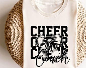 May include: Cream-coloured t-shirt featuring the text "CHEER COACH" in a black, bold font, and a detailed bow graphic. The shirt is folded and placed on a woven, circular surface.