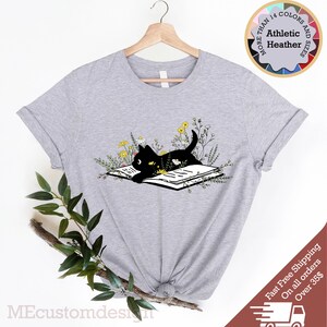 May include: A gray t-shirt with a black cat reading a book surrounded by yellow flowers. The text "MORE THAN 14 COLORS AND SIZES" is in a circle above the cat.