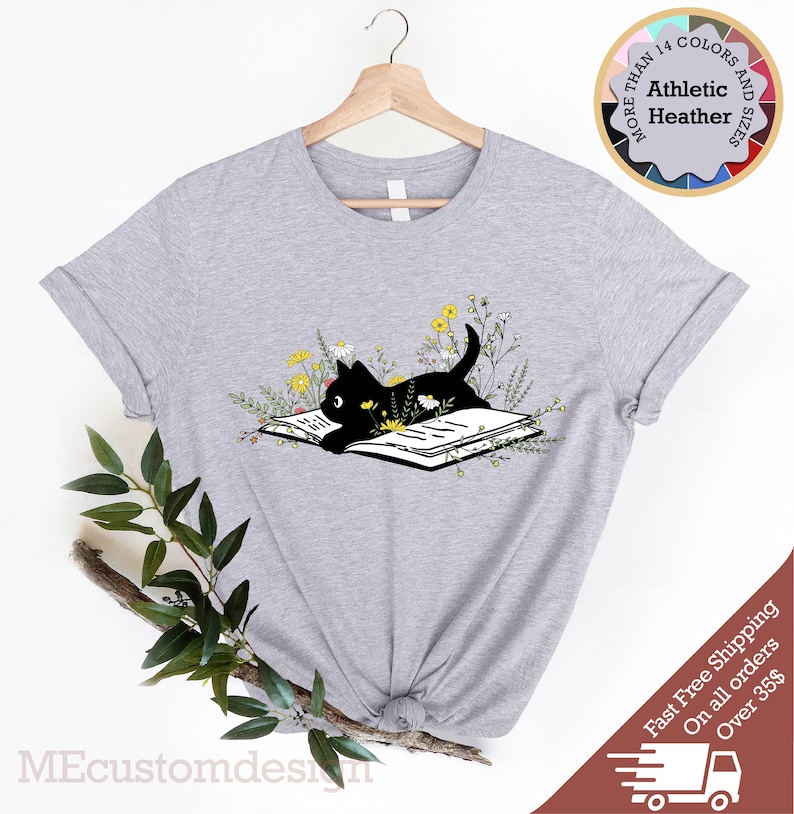 May include: A gray t-shirt with a black cat reading a book surrounded by yellow flowers. The text "MORE THAN 14 COLORS AND SIZES" is in a circle above the cat.