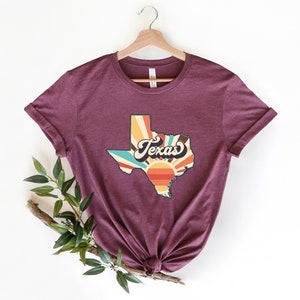 May include: A maroon t-shirt with a vintage-style graphic of the state of Texas with a sunburst design and the word "Texas" in a retro font.