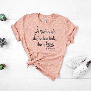 May include: A peach-colored t-shirt with the quote "And though she be but little, she is fierce" in black script. The shirt is knotted at the bottom and paired with white sneakers. Small potted succulents are arranged on a white wooden surface.