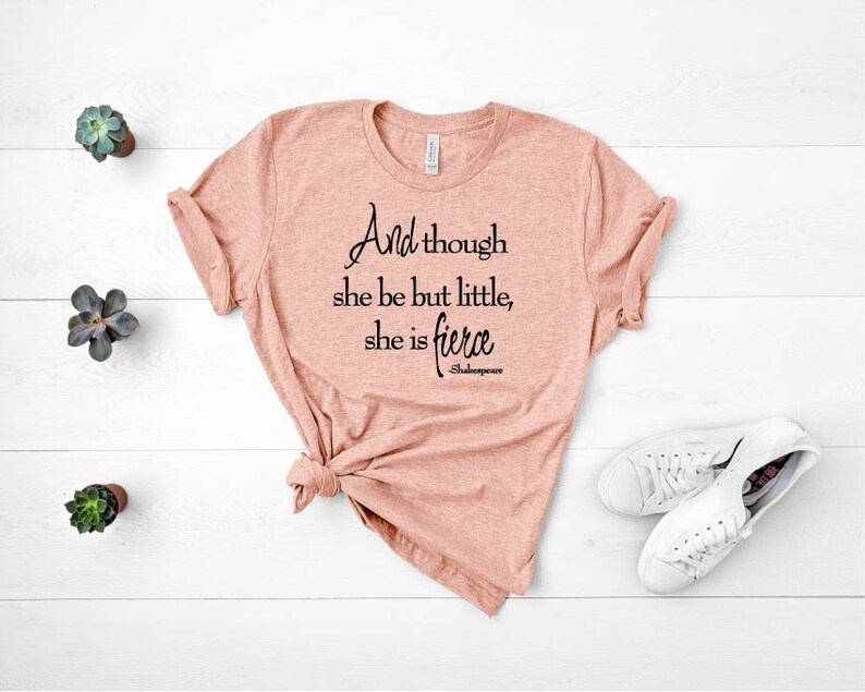 May include: A peach-colored t-shirt with the quote "And though she be but little, she is fierce" in black script. The shirt is knotted at the bottom and paired with white sneakers. Small potted succulents are arranged on a white wooden surface.