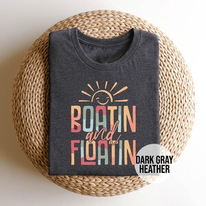 May include: Dark gray heather t-shirt with the words "BOATIN and FLOATIN" in colorful block letters, topped with a smiling sun graphic. The shirt is folded on a woven, circular surface. The words "DARK GRAY HEATHER" are in a white circle.