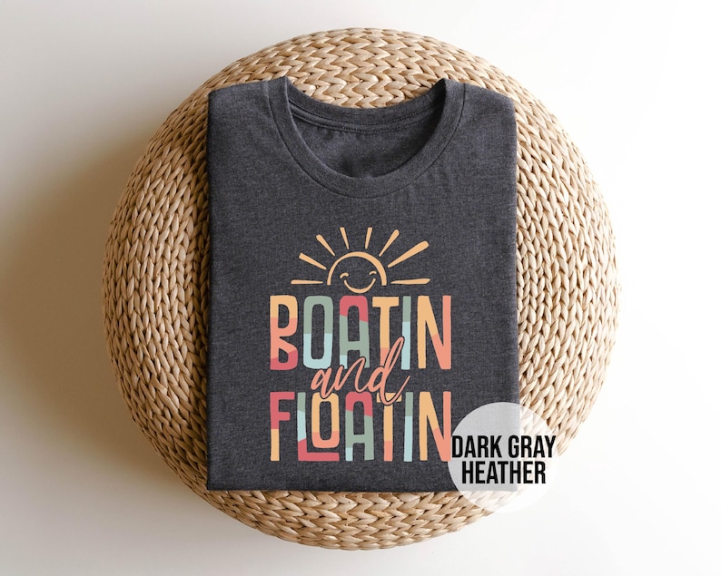May include: Dark gray heather t-shirt with the words "BOATIN and FLOATIN" in colorful block letters, topped with a smiling sun graphic. The shirt is folded on a woven, circular surface. The words "DARK GRAY HEATHER" are in a white circle.