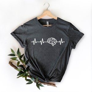Op de afbeelding: Een donkergrijs T-shirt met een witte hersengrafiek en een hartslaglijn. Het shirt hangt aan een hanger en heeft een witte achtergrond.