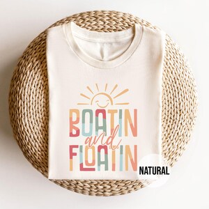 May include: Cream-colored t-shirt with the words "BOATIN and FLOATIN" in colorful, retro-style lettering, topped with a smiling sun graphic. The word "NATURAL" is printed on the bottom right.
