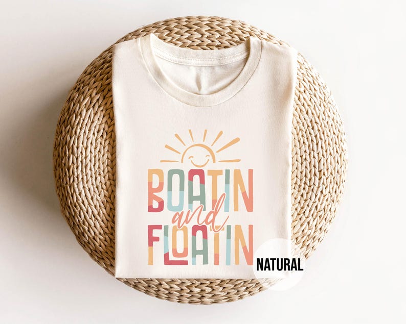 May include: Cream-colored t-shirt with the words "BOATIN and FLOATIN" in colorful, retro-style lettering, topped with a smiling sun graphic. The word "NATURAL" is printed on the bottom right.