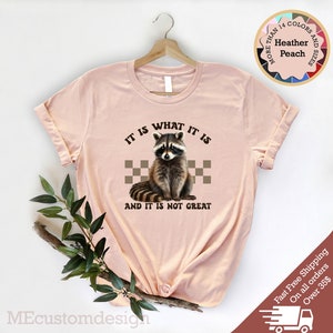 Pu&ograve; includere: Una maglietta rosa con un'immagine di un procione e il testo "IT IS WHAT IT IS AND IT IS NOT GREAT".
