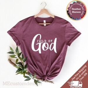 May include: A maroon t-shirt with the text "CHILD OF GOD" in white lettering. The shirt is hanging on a wooden hanger.