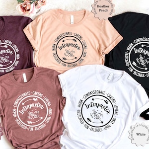 May include: Four t-shirts in different colours with a circle design that says "Warm-Compassionate-Caring-Loving-Interpreter-Dedicated-Fun-Reliable-Loyal-Kind". The shirts are heather mauve, white, black, and heather peach.