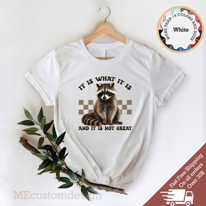 Pu&ograve; includere: Maglietta bianca con un grafico di un procione e il testo "IT IS WHAT IT IS AND IT IS NOT GREAT".