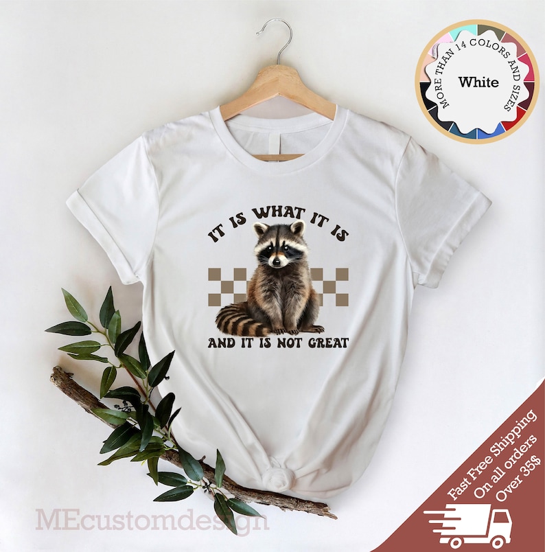 Pu&ograve; includere: Maglietta bianca con un grafico di un procione e il testo "IT IS WHAT IT IS AND IT IS NOT GREAT".