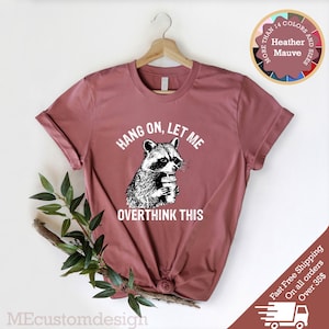 May include: A heather mauve t-shirt with a graphic of a raccoon holding a cup of coffee. The text "Hang on, let me overthink this" is printed on the shirt.