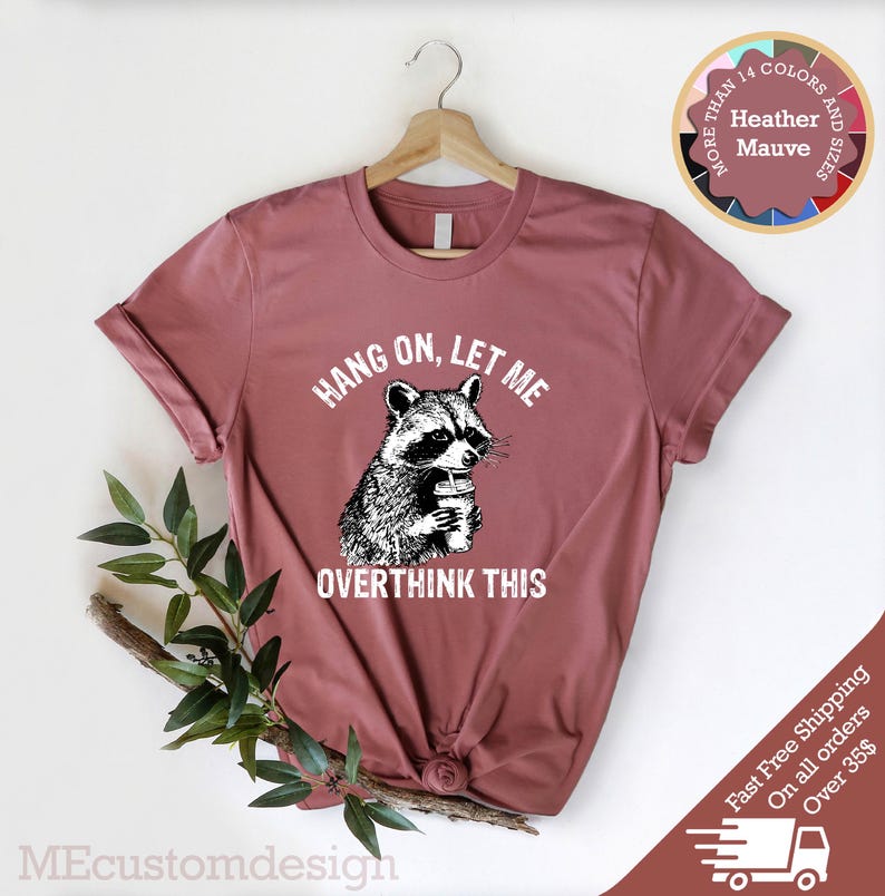 May include: A heather mauve t-shirt with a graphic of a raccoon holding a cup of coffee. The text "Hang on, let me overthink this" is printed on the shirt.