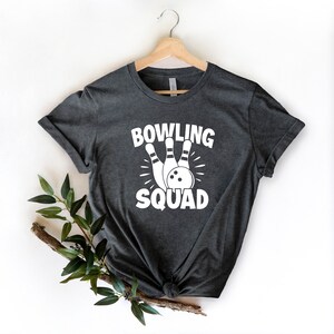May include: A dark gray t-shirt with a white graphic of bowling pins and a bowling ball. The text "BOWLING SQUAD" is printed on the shirt.