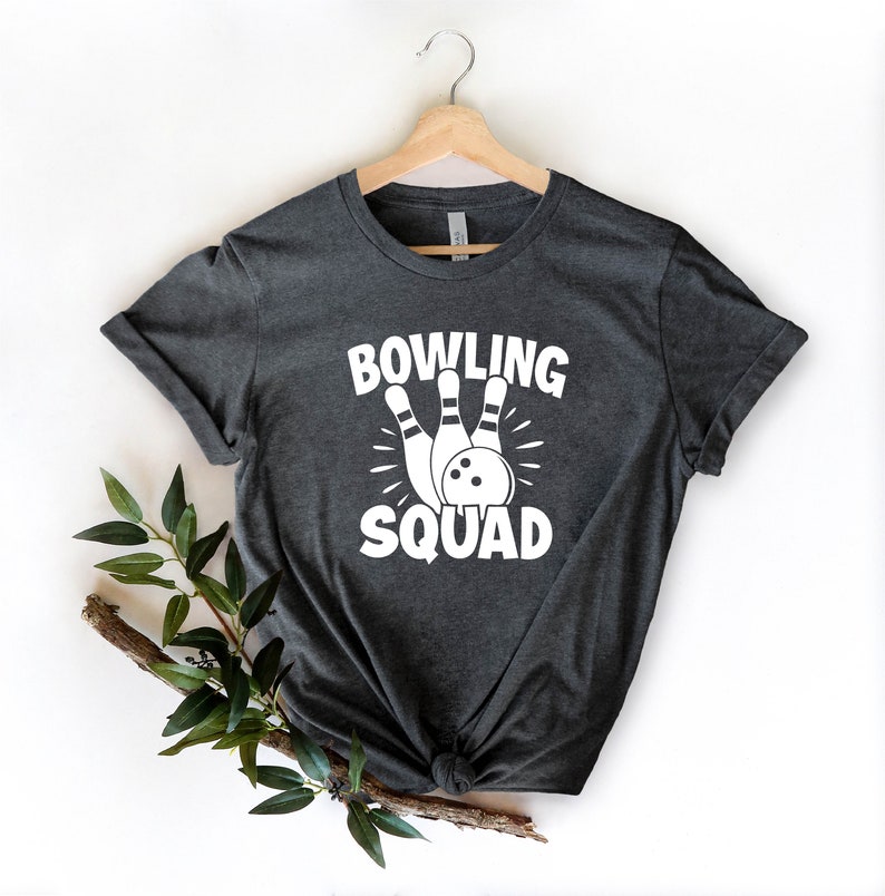 May include: A dark gray t-shirt with a white graphic of bowling pins and a bowling ball. The text "BOWLING SQUAD" is printed on the shirt.