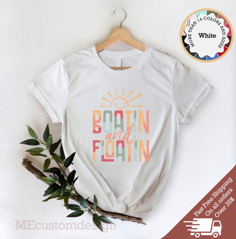 May include: White t-shirt with the words "BOATING and FLOATING" in colorful block letters, with a smiling sun graphic above. The shirt is displayed on a wooden hanger.