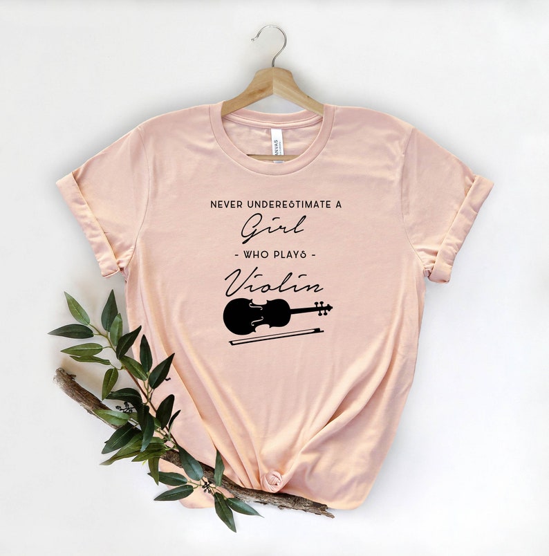 May include: A light pink t-shirt with the text "Never underestimate a girl who plays violin" in black lettering. A black silhouette of a violin and bow is below the text.