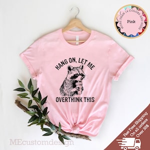 May include: Pink t-shirt with a black and white illustration of a raccoon holding a coffee cup. The text "HANG ON, LET ME OVERTHINK THIS" is printed on the shirt.