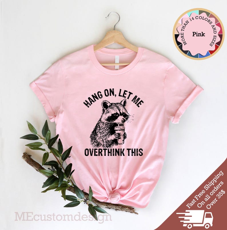 May include: Pink t-shirt with a black and white illustration of a raccoon holding a coffee cup. The text "HANG ON, LET ME OVERTHINK THIS" is printed on the shirt.