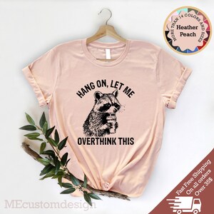 May include: A light pink t-shirt with a black graphic of a raccoon holding a cup and the text "Hang on, let me overthink this".