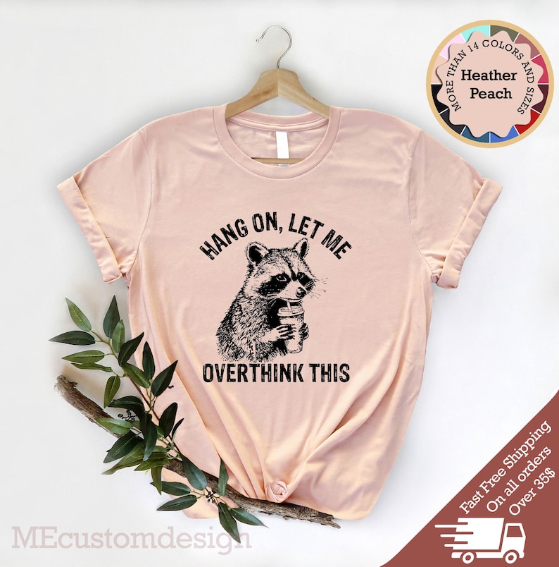 May include: A light pink t-shirt with a black graphic of a raccoon holding a cup and the text "Hang on, let me overthink this".