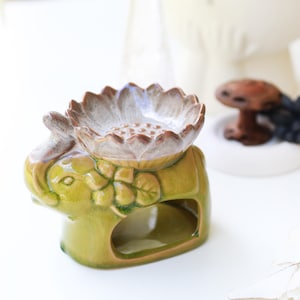 May include: A ceramic oil burner shaped like a green elephant with a glossy finish. The top of the elephant features a wide, shallow bowl designed like a lotus flower with a light beige and brown gradient glaze, resting on the elephant's back.